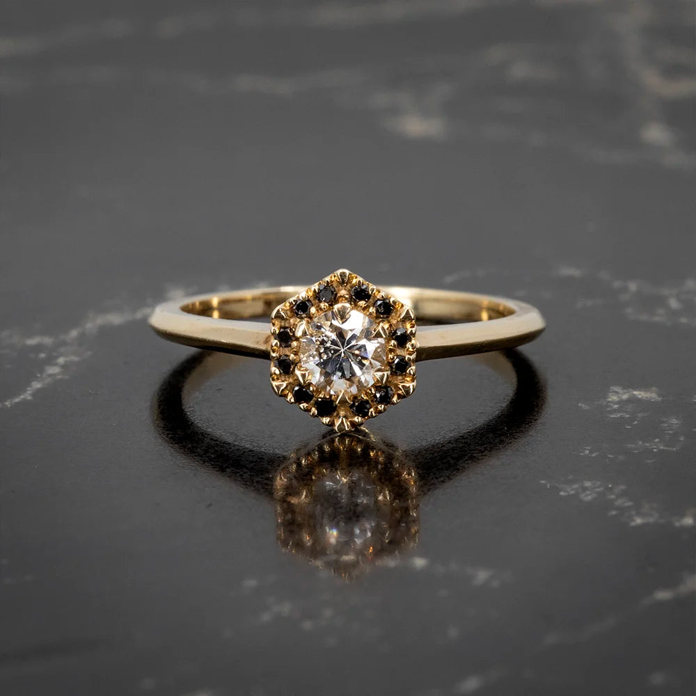 Whimsical engagement ring by Liane Vaz, featuring a brilliant-cut diamond framed by a halo of black diamonds in a hexagonal design. Available at Ruby Mardi, Montreal.