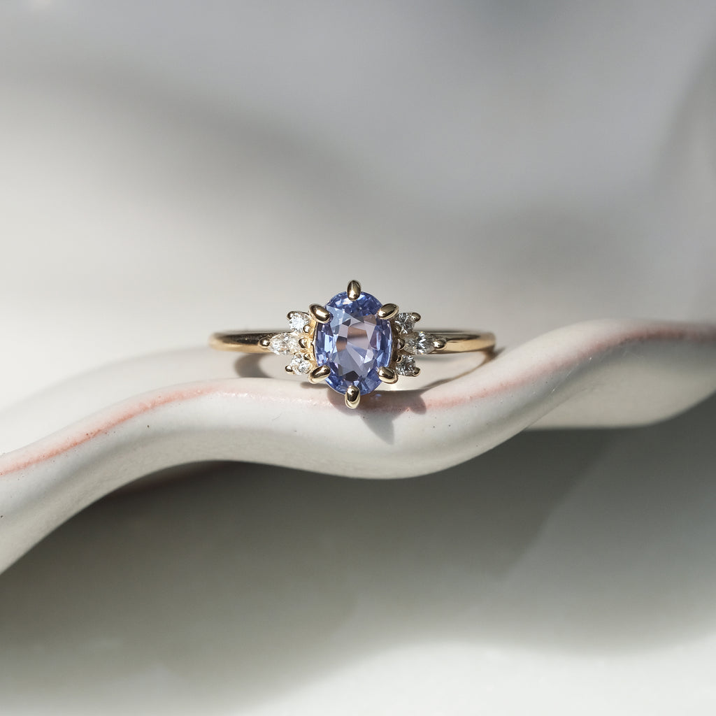 Light blue sapphire and diamond engagement ring in 14k yellow gold, displayed on a ceramic surface. Designed by Maksym Jewelry, available at Ruby Mardi in Montreal, Canada.