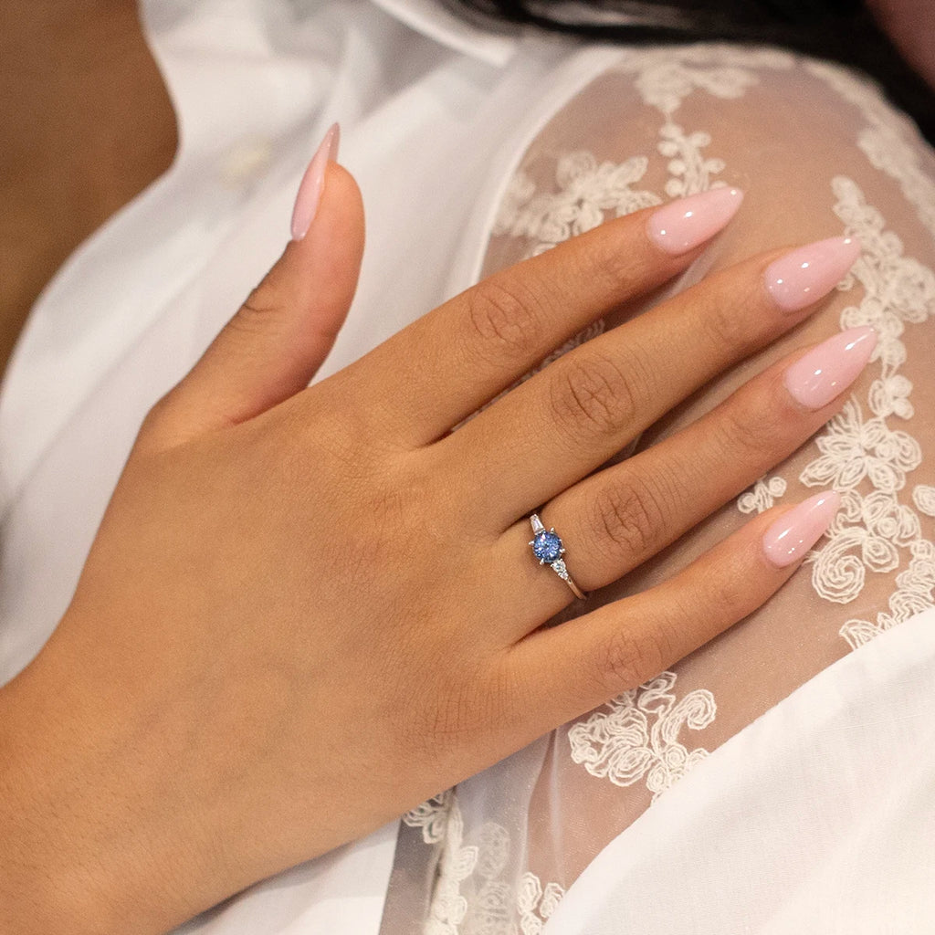 A chic hand with light pink nails wears a beautiful asymmetrical engagement ring with a central round blue sapphire and diamond accents. One-of-a-kind creation available worldwide through at Ruby Mardi's website.