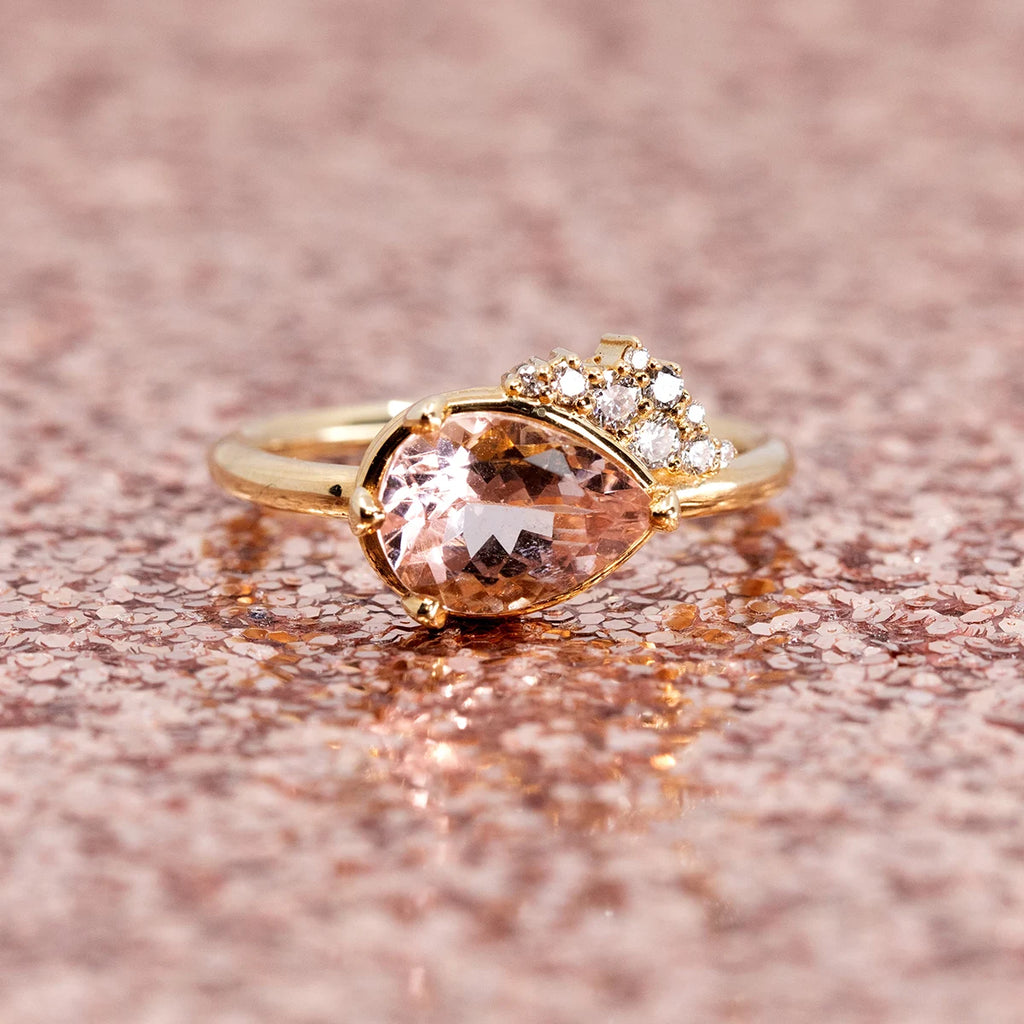 A stunning pear-shaped morganite engagement ring set in 14k yellow gold, accented with a cluster of sparkling diamonds. Handcrafted by Sheena Purcell and photographed on a shimmering rose gold sequin background, available at Ruby Mardi in Montreal.