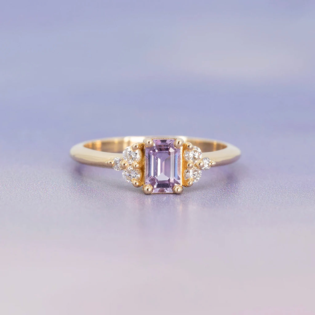 Light purple sapphire engagement ring with emerald-cut center stone and round diamonds on each side, set in yellow gold. Fine jewelry by Ruby Mardi, Montreal.
