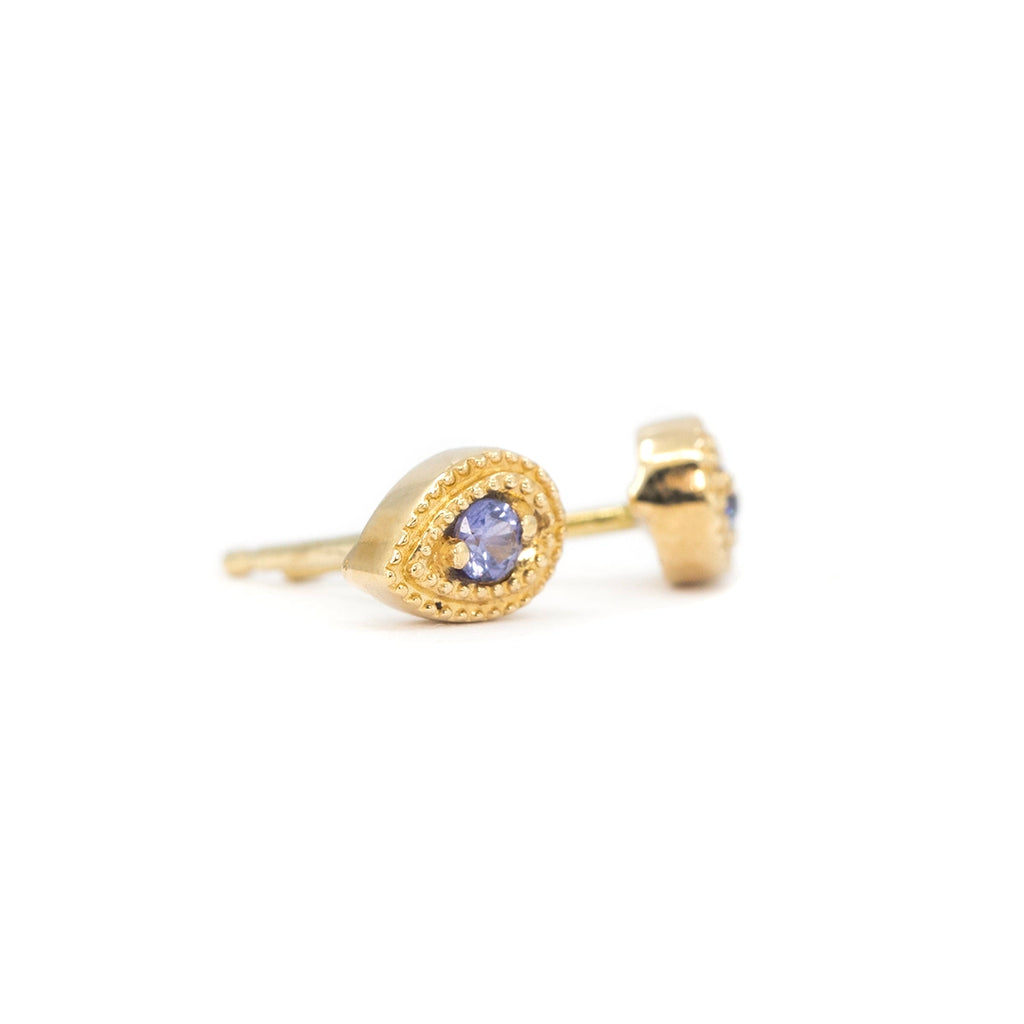 Close-up of Emily Gill’s stud earrings in 14k yellow gold, featuring round lilac-purple sapphires set in teardrop frames with delicate milgrain beading. Minimalist yet vintage-inspired gemstone earrings, handcrafted designer jewelry available at Ruby Mardi in Montreal.