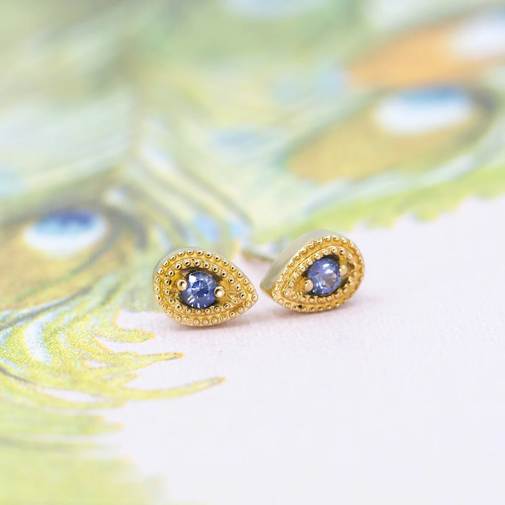 Pair of teardrop stud earrings in 14k yellow gold by Emily Gill, each set with a round lilac-purple sapphire and framed with intricate milgrain beading. Handcrafted gemstone jewelry with vintage-inspired charm, photographed on a soft peacock feather background. Available at Ruby Mardi, Montreal jewelry store.
