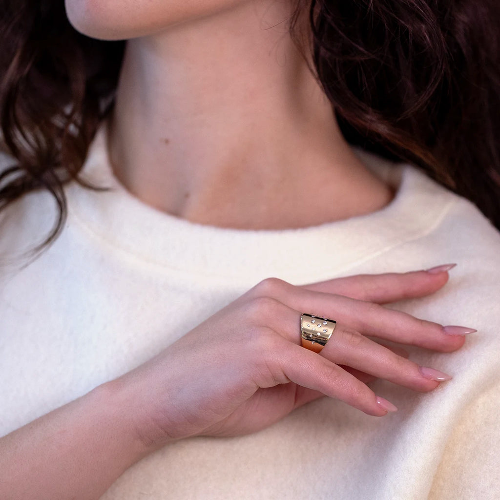 A close-up of a woman's hand wearing a bold yellow gold ring with scattered diamonds. The ring has a sculptural design and is worn on the ring finger. The woman is dressed in a soft white sweater, and her dark hair cascades down her shoulder. Available through Ruby Mardi, a unique collective of indie designer jewelry in Canada.