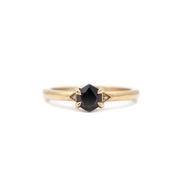 Close-up product image of the Luna Hex ring by Liane Vaz, featuring a hexagon-cut black spinel center stone flanked by two tiny champagne diamonds, set in a polished yellow gold band. The ring is photographed against a plain white background for Ruby Mardi jewelry in Montreal, Canada.