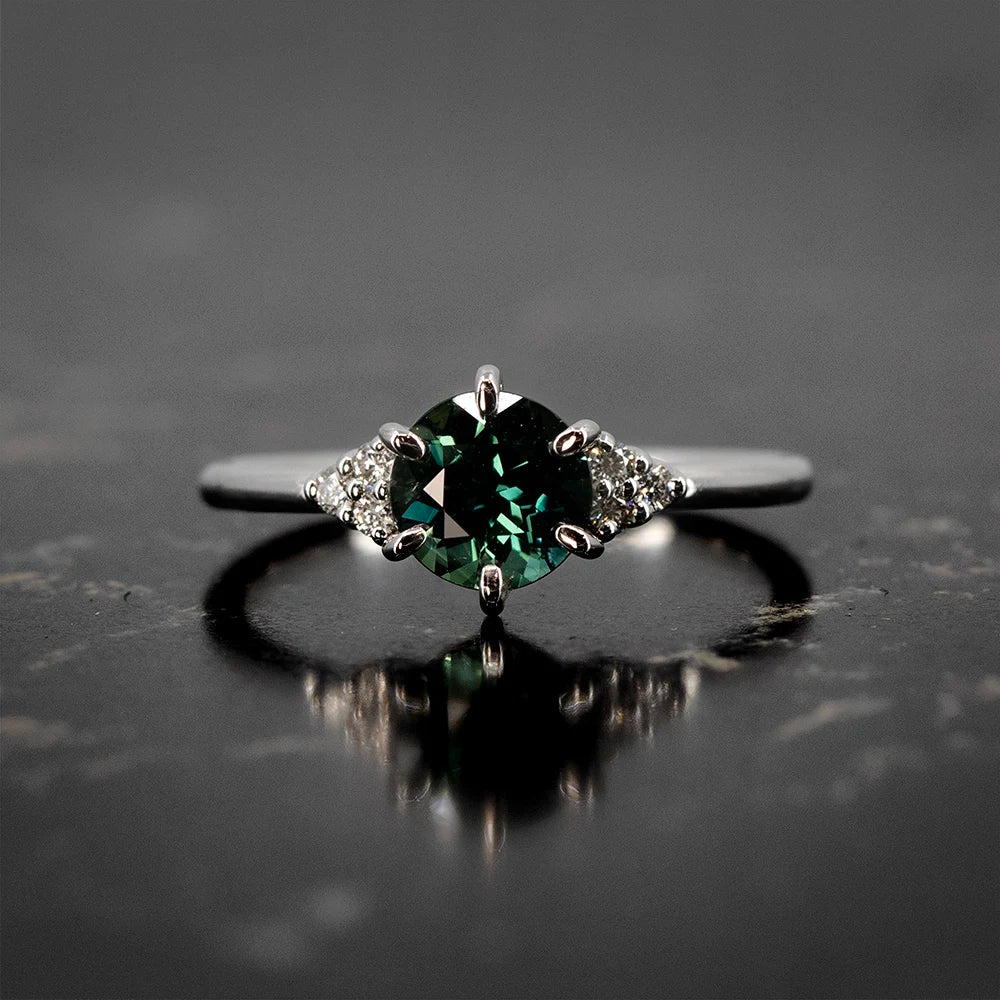 White gold engagement ring with a round dark green sapphire center stone and diamond cluster accents by Maksym Jewelry, available at Ruby Mardi.