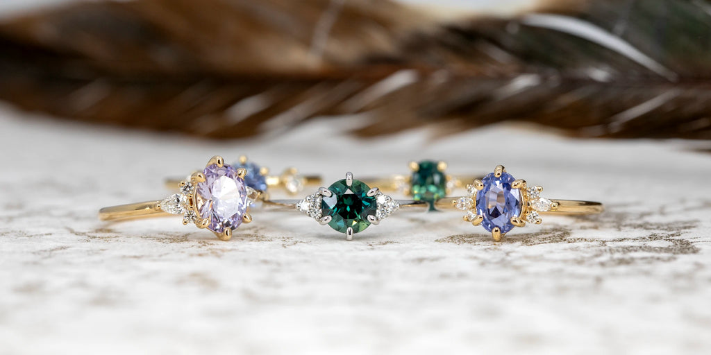 Collection of Maksym Jewelry rings with oval and round sapphires in various colors, yellow and white gold bands with diamond accents, displayed on textured surface.