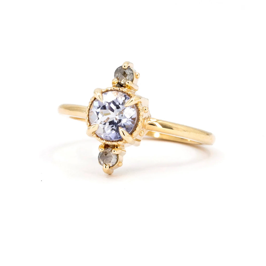 Unique, alternative engagement ring photographed from the front against a white background. Details of gold granules can be seen on the sides of the ring. The fine jewel is bezel-set with eagle's paw claws. It features a splendid rose-cut lavender sapphire and salt-and-pepper diamond accents. It's from Nadia Werchola and available at independent jewelry store Ruby Mardi.
