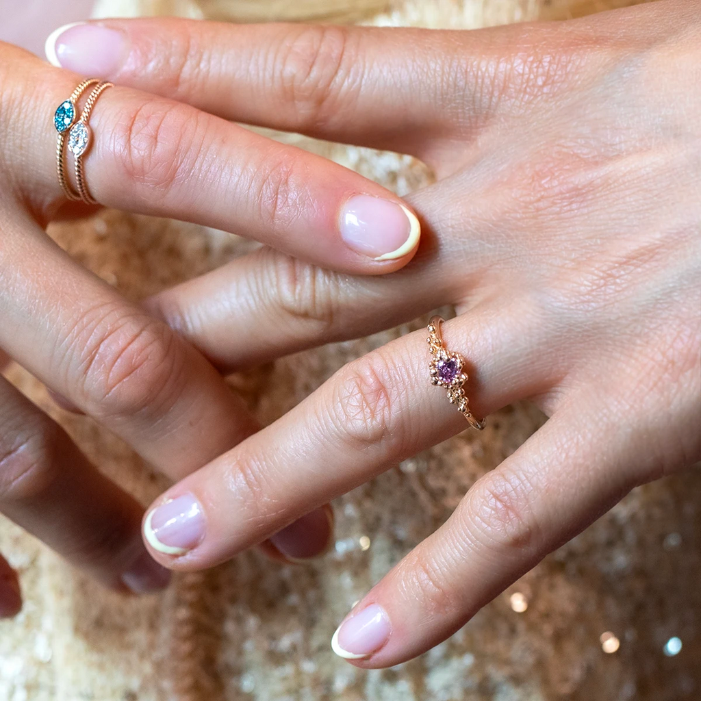 Close-up of a hand wearing two gold rings: one with teal and white gemstones, and another solitaire engagement ring with a purple sapphire and organic band. Designer jewelry by Ruby Mardi, Montreal, Canada.