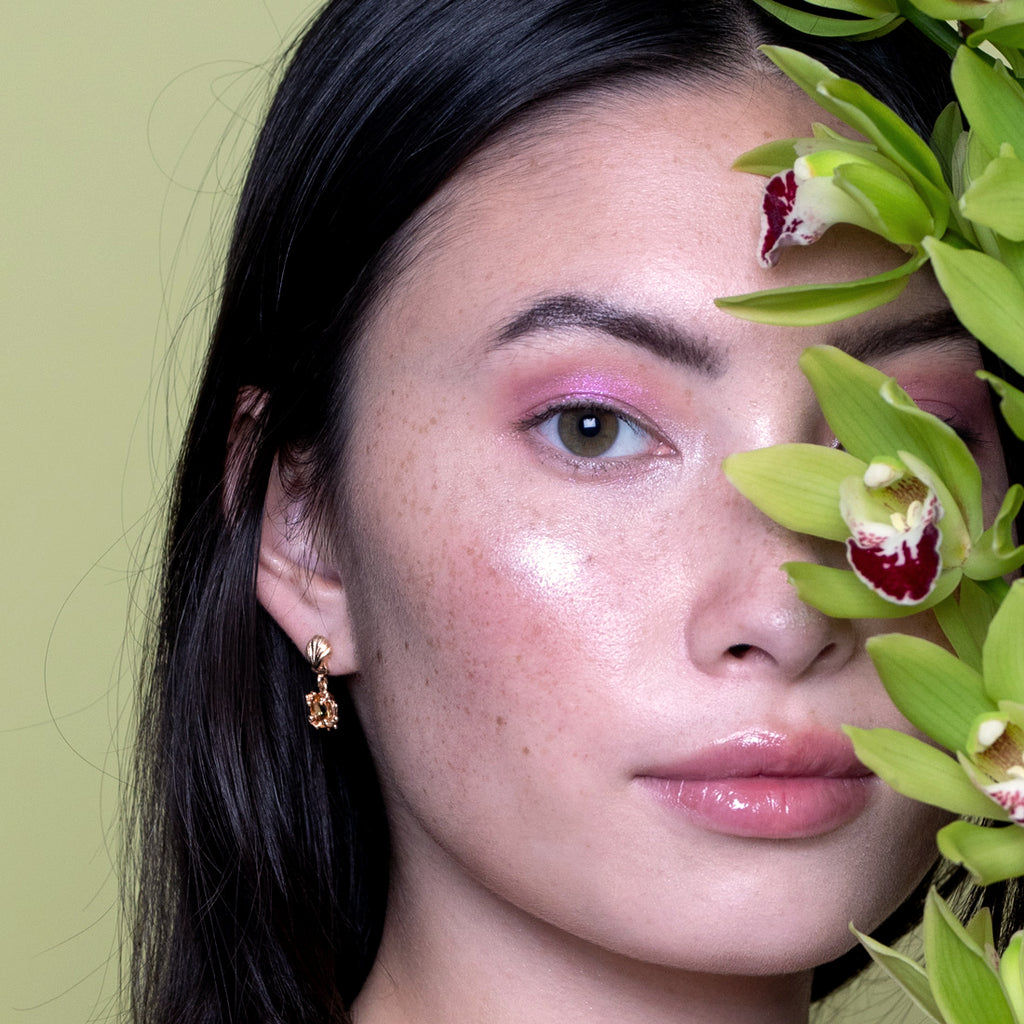 Close-up portrait of a woman wearing yellow gold shell earrings with yellow sapphire, designed by Meg Lizabet. Artistic designer jewelry from Ruby Mardi, photographed with green orchids in the background.
