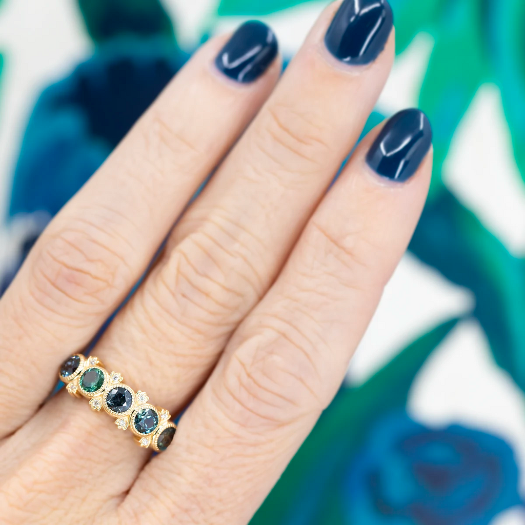 We see in close up 3 fingers with dark blue nails photographed in front of an abstract blue and green background. On the middle finger, we can see a large sapphire band that presents teal round sapphires bezel set with miligrain details. There are also 8 small natural diamond accents that enhance the composition. This one-of-a-kind designer ring is available at fine jewellery store Ruby Mardi in Montreal, Canada.