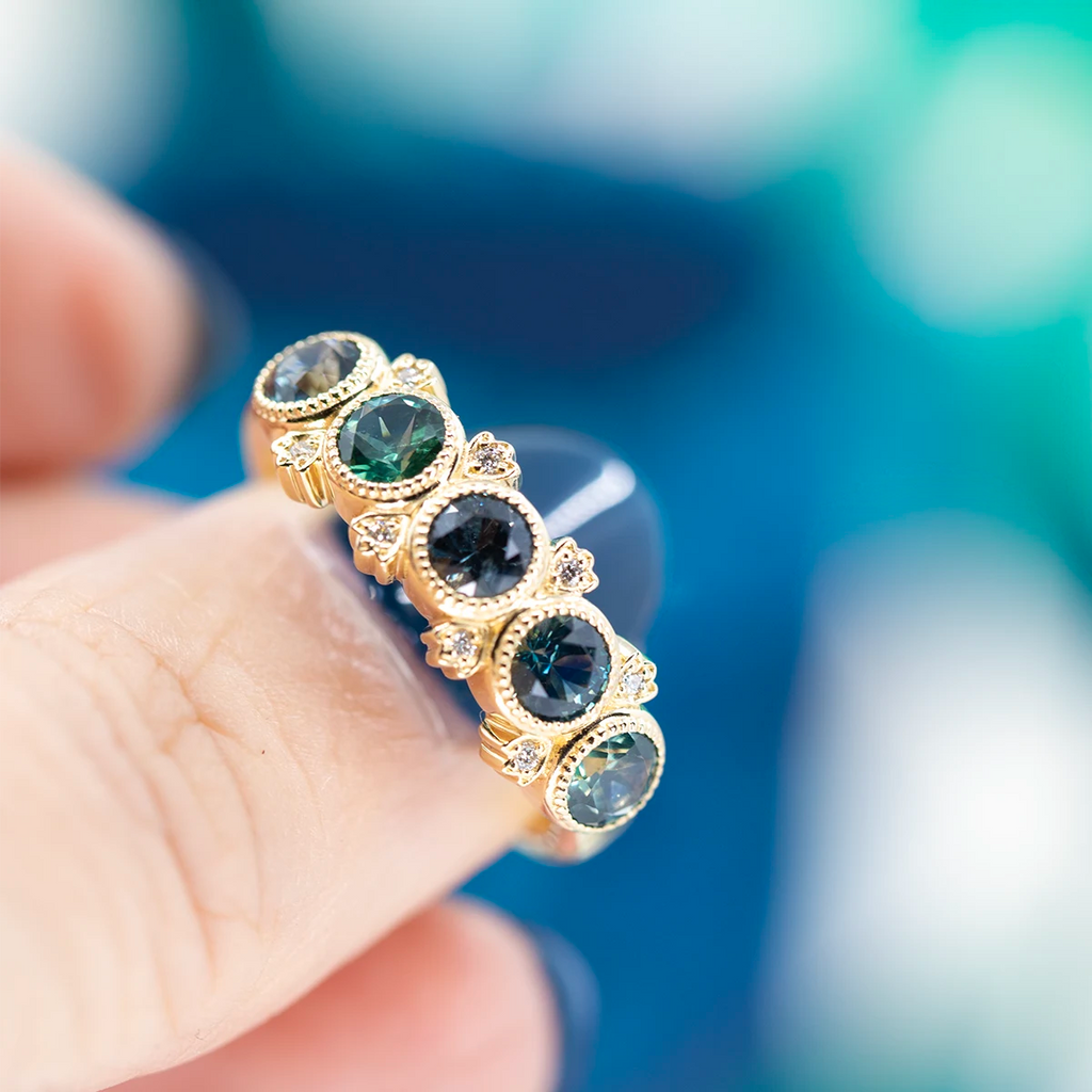 Close-up photo of a ring on the tip of a thumb with a dark-blue nail: the ring features 5 large, round, ethically-sourced natural sapphires in shades of blue-green, bezel set with miligrain details and 8 small, natural white accent diamonds.