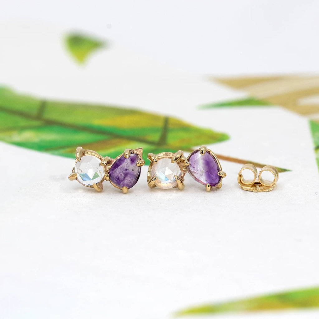 One-of-a-kind 10k yellow gold stud earrings by Toronto independent artist Nadia Werchola, featuring natural amethysts and rose-cut moonstones in slightly irregular eagle-claw prong settings, photographed on a white surface with soft green botanical details, available at Ruby Mardi in Montreal.