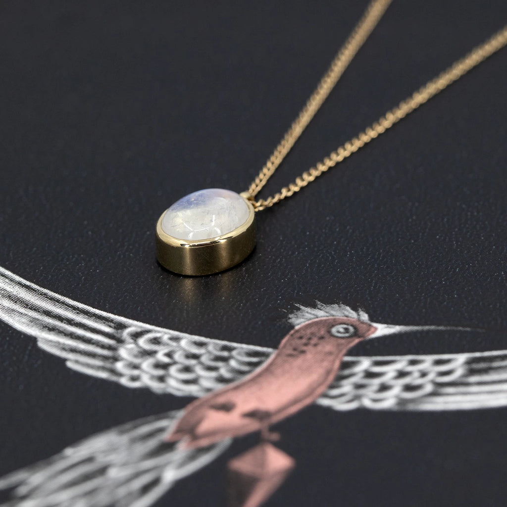Flat lay of a 10k yellow gold pendant with an oval cabochon moonstone, bezel set, photographed on a dark background with a delicate pink and white bird illustration. This side view captures the pendant's volume and fine craftsmanship, designed by Rooz Jewelry and available at Ruby Mardi.