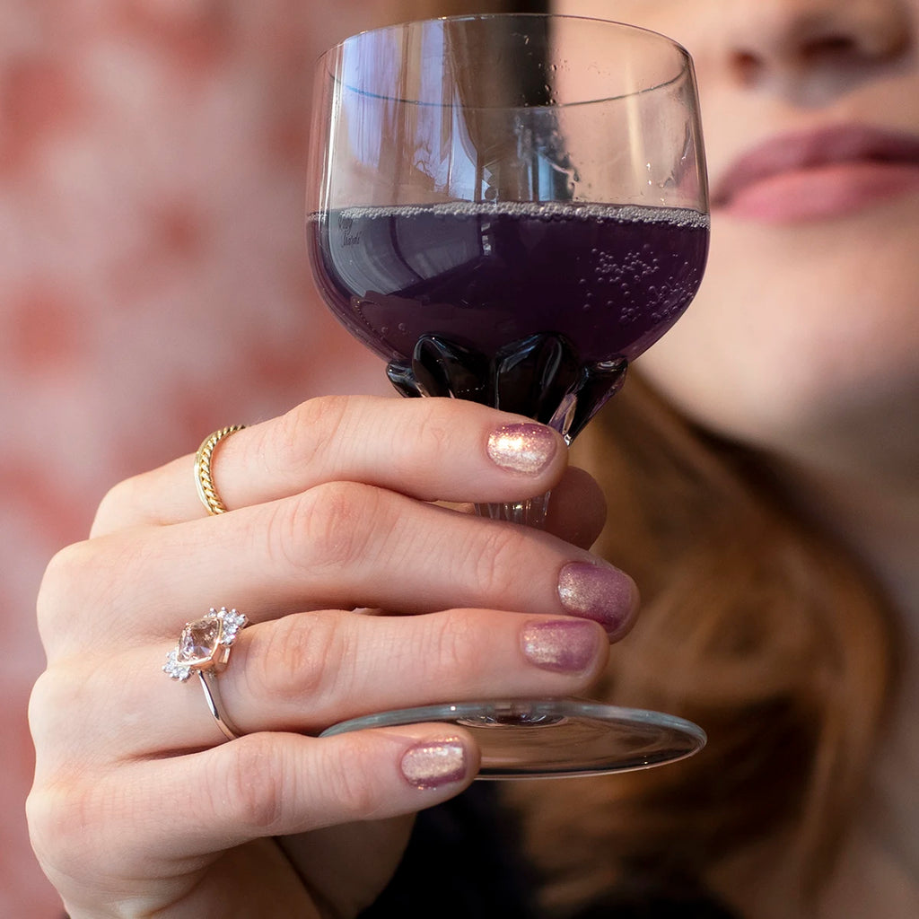 A young woman photographed close-up holds up a cup containing a violet liquid. On her hand are gold rings, including, in the foreground, a stunning rose and white gold engagement ring with cushion-shaped morganite and asymmetrically placed natural diamond accents.