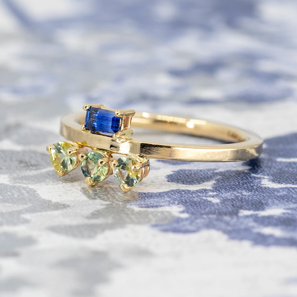 Fancy engagement ring with 4 sapphires set in 14k recycled gold. There's on top blue sapphire baguette, and 3 round parti yellow-green sapphires on the bottom of the band. This unique engagement ring can be worn in both ways. It's a design from independent brand In the Light of Day Jewelry, and it is sold by Ruby Mardi, a funky jewelry store in Montreal, Canada. 