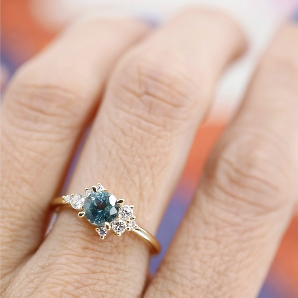 Charming product photography of a one-of-a-kind engagement ring that dazzles with a stunning round soft blue sapphire at its center, surrounded by a brilliant cluster of natural diamonds. Worn gracefully on a finger, this exquisite piece showcases its unique asymmetrical design and exceptional craftsmanship. A true masterpiece by Justine Quintal, now available at Ruby Mardi, Canada’s premier jewelry destination.
