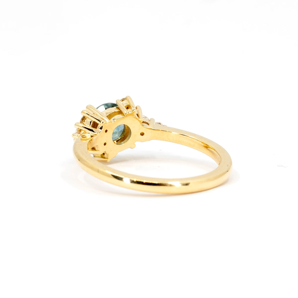 Elegant product photography of a unique engagement ring featuring a stunning round soft blue sapphire and a cluster of natural diamonds, beautifully displayed against a white background. This captivating shot highlights the intricate detailing and craftsmanship of the ring's back design, showcasing the care and artistry that goes into each piece. A stunning creation by Justine Quintal, available exclusively at Ruby Mardi, Canada's finest jewelry store.
