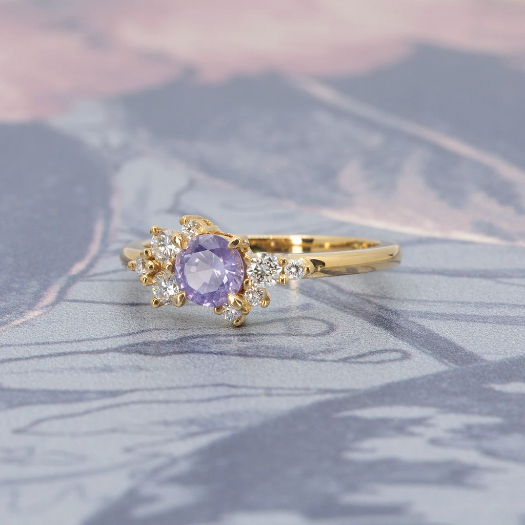 A gold ring handcrafted by Justine Quintal, featuring a round purple sapphire center stone with asymmetrically arranged small round white diamond accents, displayed on a soft-focus patterned background in shades of blue and pink.