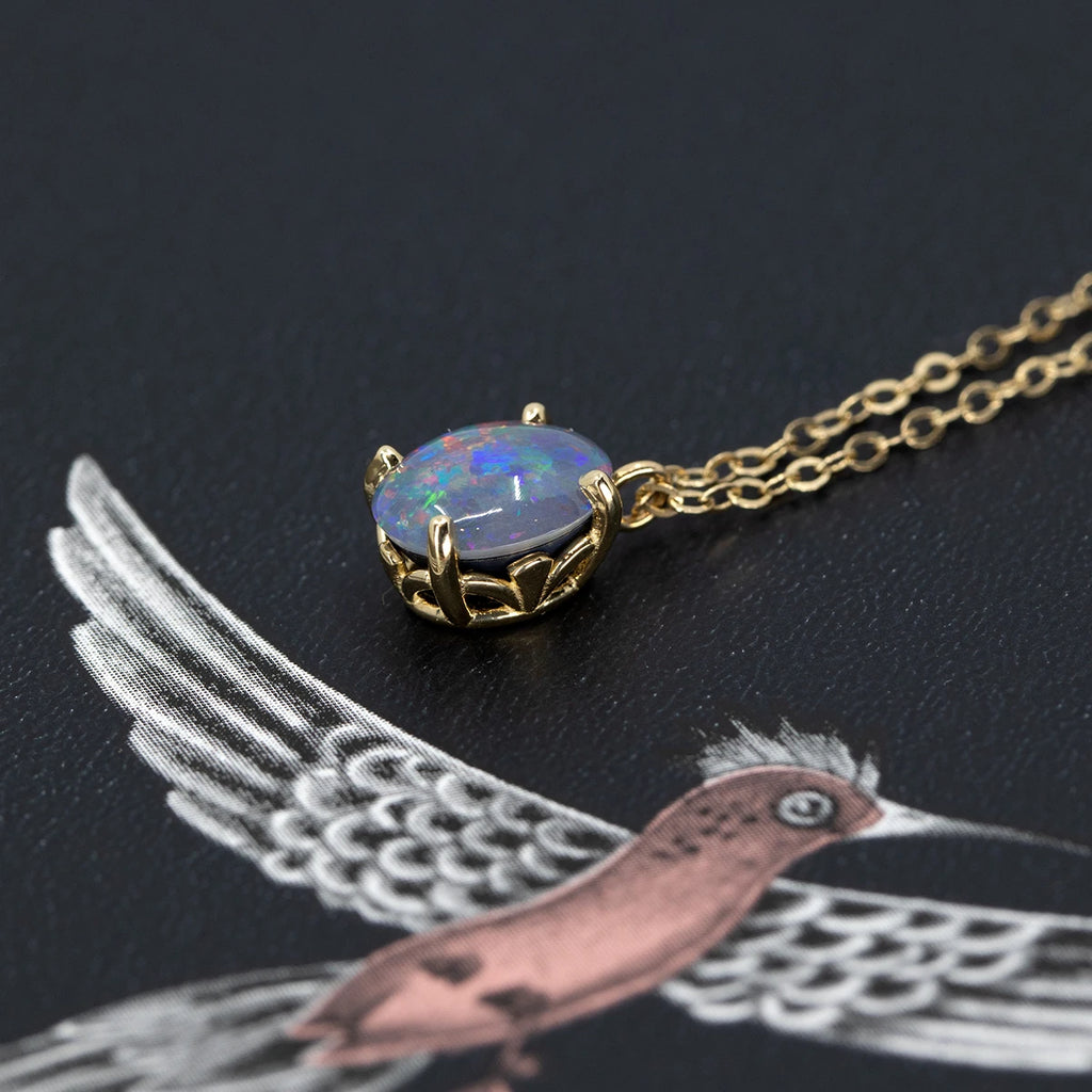 A beautiful natural blue opal set in a gold basket with feminine details is seen photographed on a cool background with a white and pink bird. This designer jewelry piece is one-of-a-kind and only available at Ruby Mardi, a Montreal jewelry store that ships worldwide.