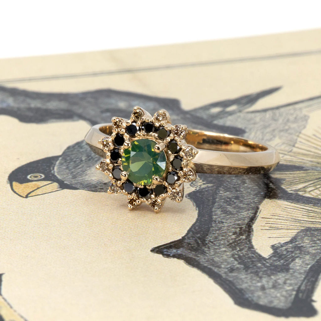 Yellow gold ring with a round green opalescent sapphire, surrounded by a double halo of black and white diamonds in a starburst pattern. Photographed on a vintage bird illustration background. Handcrafted in Montreal by Ruby Mardi.