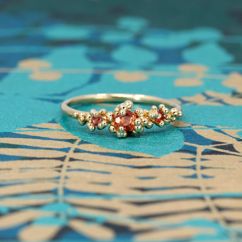 Gold ring with three orange sapphires surrounded by organic gold bead accents, designed as an alternative engagement ring. Part of the Areia Signature Collection by Meg Lizabet for Ruby Mardi. Photographed on a vibrant blue botanical background. Handcrafted in Montreal, Canada.