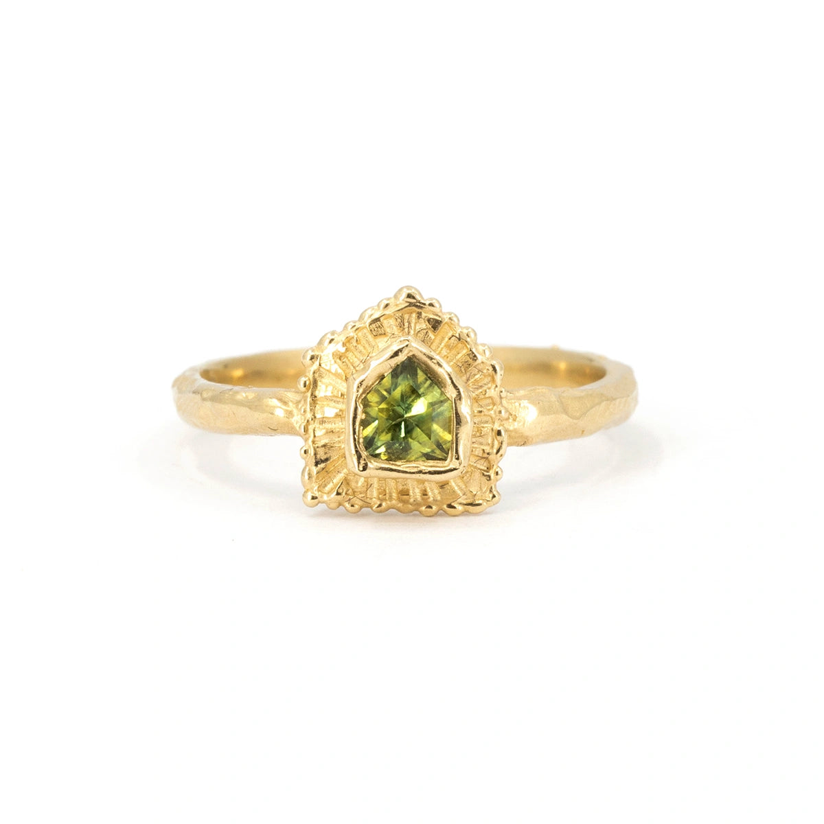 ‘Coming Home’ ring by Emma Glover, exclusive to Ruby Mardi. Crafted in 10k yellow gold, it features a green sapphire with yellow hues, shaped like a house. A hand-carved gold halo surrounds the stone, enhancing its house-like appearance. The thick, irregular gold band highlights its organic, handcrafted design. Seen on a white background.