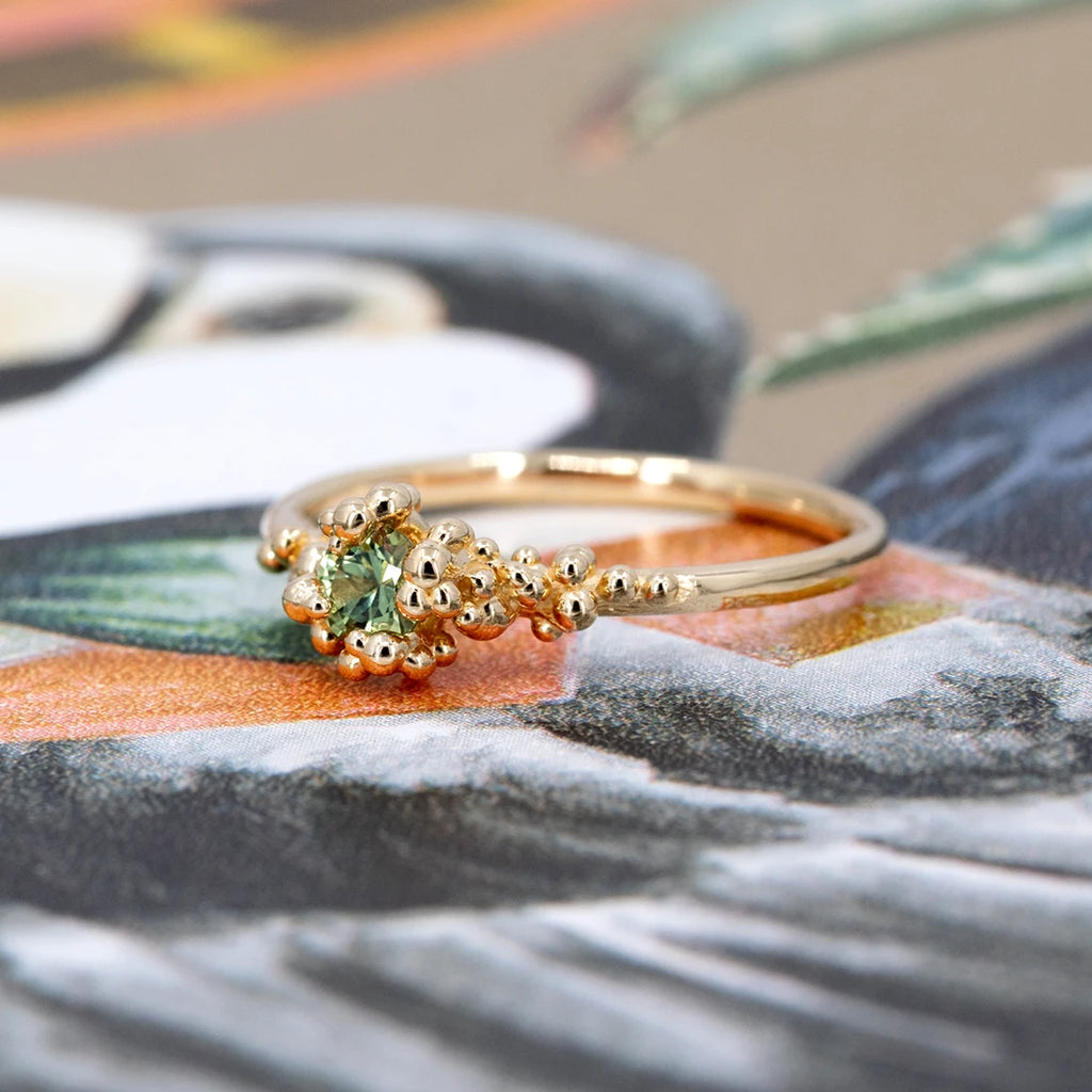 Organic 14k gold engagement ring with light green sapphire and sculptural granules, handmade by Meg Lizabet. Artistic jewelry shot on a colorful illustration background, available at Ruby Mardi, Montreal.