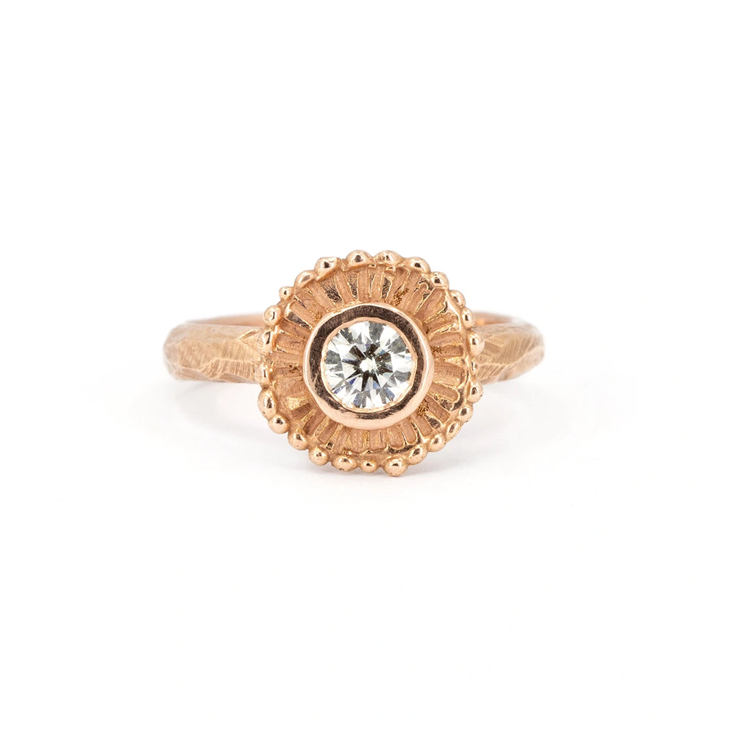 Front view on a white background of a fairly solid rose gold jewel with a raw or “organic” style. The band itself is wide and textured. In the center, a natural round white diamond in a closed setting, around which the rose gold forms a sunburst “halo”. This jewel is available only from Ruby Mardi, a luxury jewelry store that, like Gem Breakfast, sells the work of independent jewellery designers. 