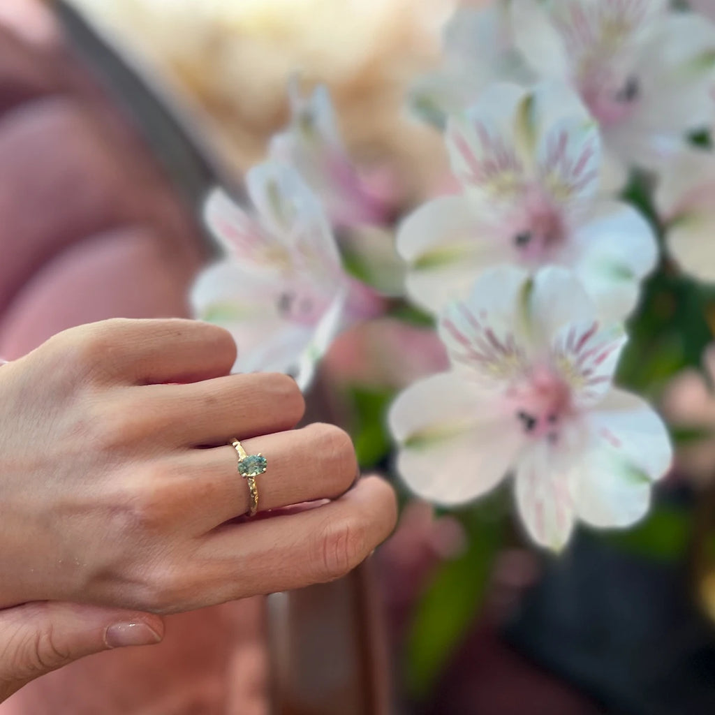 A picture showing a yellow gold ring with a green gemstone worn on a lady's hand. On the back we can see blurry white and pink flowers (alstromerias). This picture as a romantic vibe, like the solitaire engagement ring that is shown on the image.