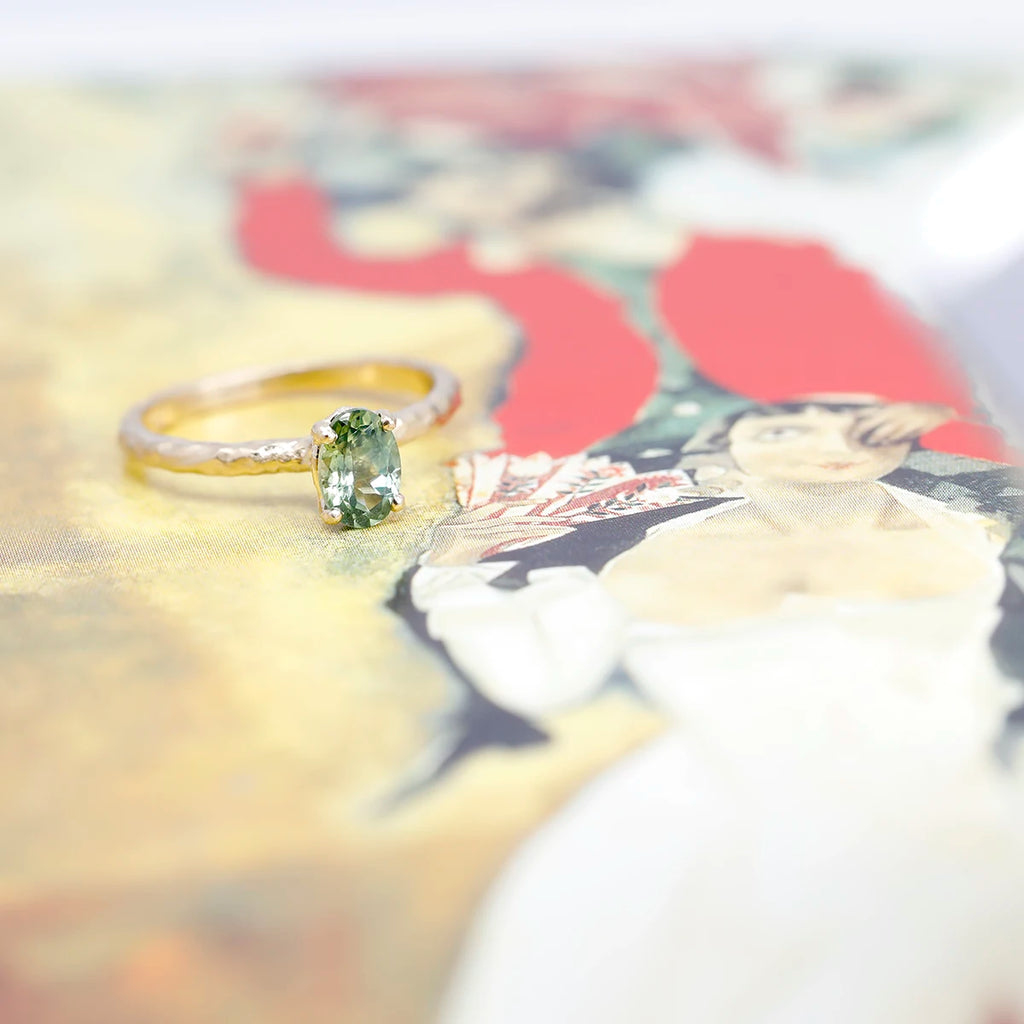 A ring with a textured band and a beautiful oval green sapphire is seen photographed on a painting showing an elegant lady. This solitaire ring was handmade in Toronto by independent jeweller Anouk Jewelry.