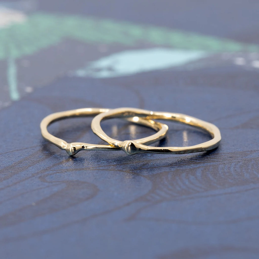 Pair of delicate 14k yellow gold rings with organic sculpted texture, handcrafted by Fluid Jewellery in Ottawa using Fairmined gold. Photographed on a blue botanical background.