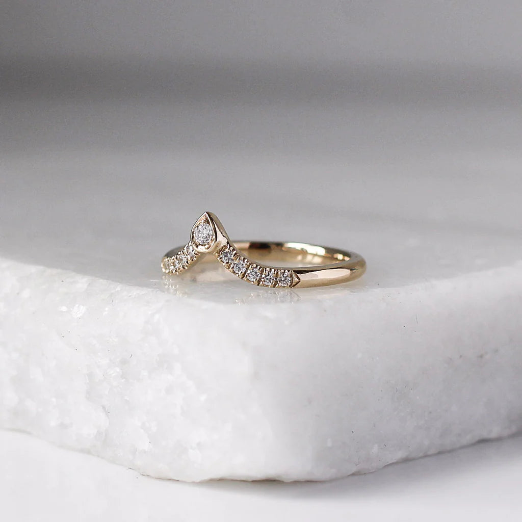 14k yellow gold diamond wave band by Toronto designer Yuliya Chorna, displayed on a white marble surface. Elegant chevron-style ring with pavé-set diamonds and a pear-shaped gold detail holding a round diamond at the peak. A modern yet timeless wedding band that complements engagement rings or can be worn alone as a refined everyday piece.