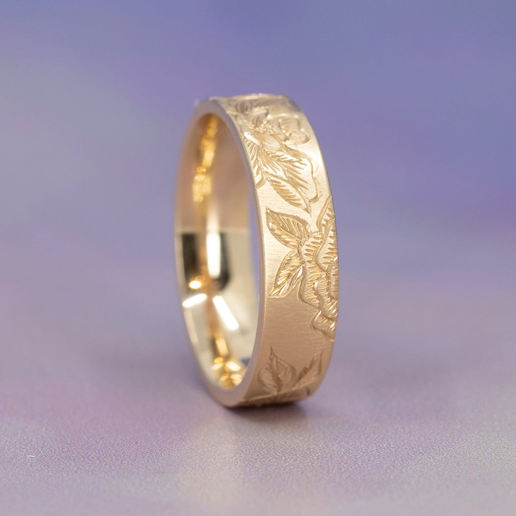Hand-engraved 14k gold band decorated with layered peony blossoms and foliage, resting on a pale surface. The ring’s soft matte texture contrasts with the bright interior, highlighting Rebel & Rogue’s artisanal floral engraving, available at Ruby Mardi.