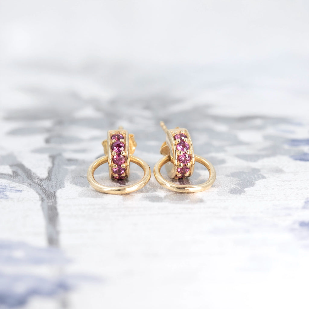 Golden hoop earrings with pink garnets by Meg Lizabet, designer jewelry at Ruby Mardi, Montreal Canada, displayed on a soft floral background.