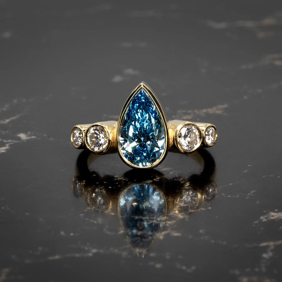 Blue diamond engagement ring with pear-shaped center stone and bezel-set side diamonds in yellow gold, crafted by Pompei Fine Jewellery and photographed on a dark marble surface. Available at Ruby Mardi, Montreal.