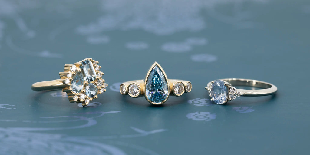 Trio of fine rings by Pompei Fine Jewellery on teal background: one yellow gold ring with aquamarine cluster and diamonds, one yellow gold ring with pear-shaped blue diamond and round diamonds, and one white gold ring with oval light sapphire and diamond accents. Handmade in Canada, available at Ruby Mardi.