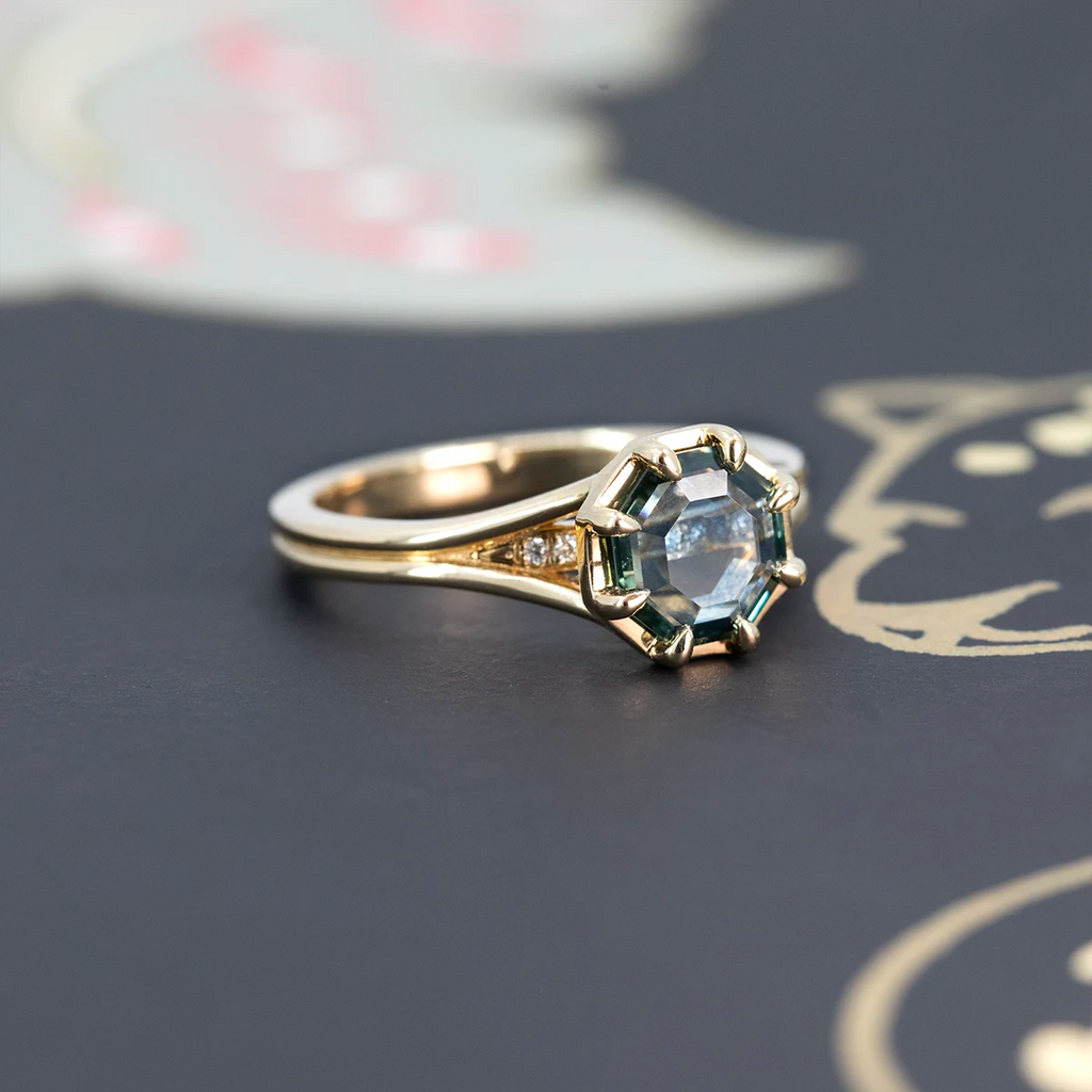 Arden ring by Emily Gill featuring a portrait-cut octagonal Montana sapphire and diamond accents, set in 14k yellow gold with a split shank, displayed on a dark illustrated background. Fine designer jewelry available at Ruby Mardi, Montreal.
