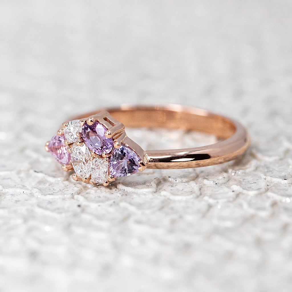 A 14k rose gold cluster ring featuring three light purple sapphires and three lab-grown diamonds, photographed from the side on a textured white snake-skin background. The unique surface enhances the ring’s romantic and vintage-inspired aesthetic.