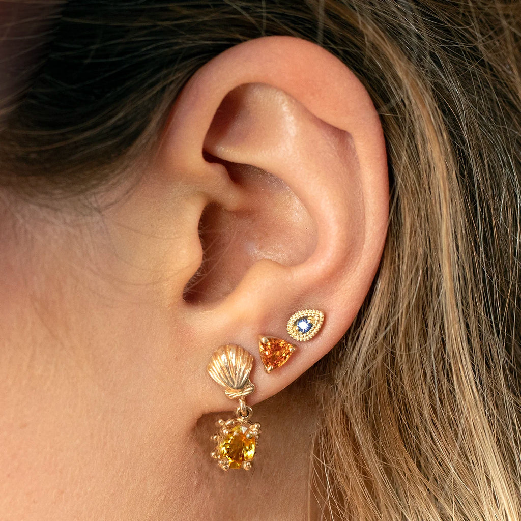 Close-up of a woman's ear wearing a stack of gold earrings, including Emily Gill’s purple sapphire stud earrings in 14k yellow gold with milgrain detail. The dainty sapphire studs are paired with bold gemstone and seashell-inspired earrings for a unique layered look. Modern designer jewelry available at Ruby Mardi in Montreal.