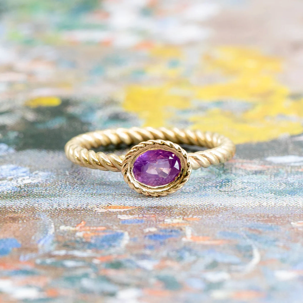 Raspberry Jam Ring by In the Light of Day Jewelry — handcrafted in 14k yellow gold with a twisted rope band and bezel-set opalescent pink sapphire, photographed on an impressionist-style painted background. Available at Ruby Mardi, Montreal.