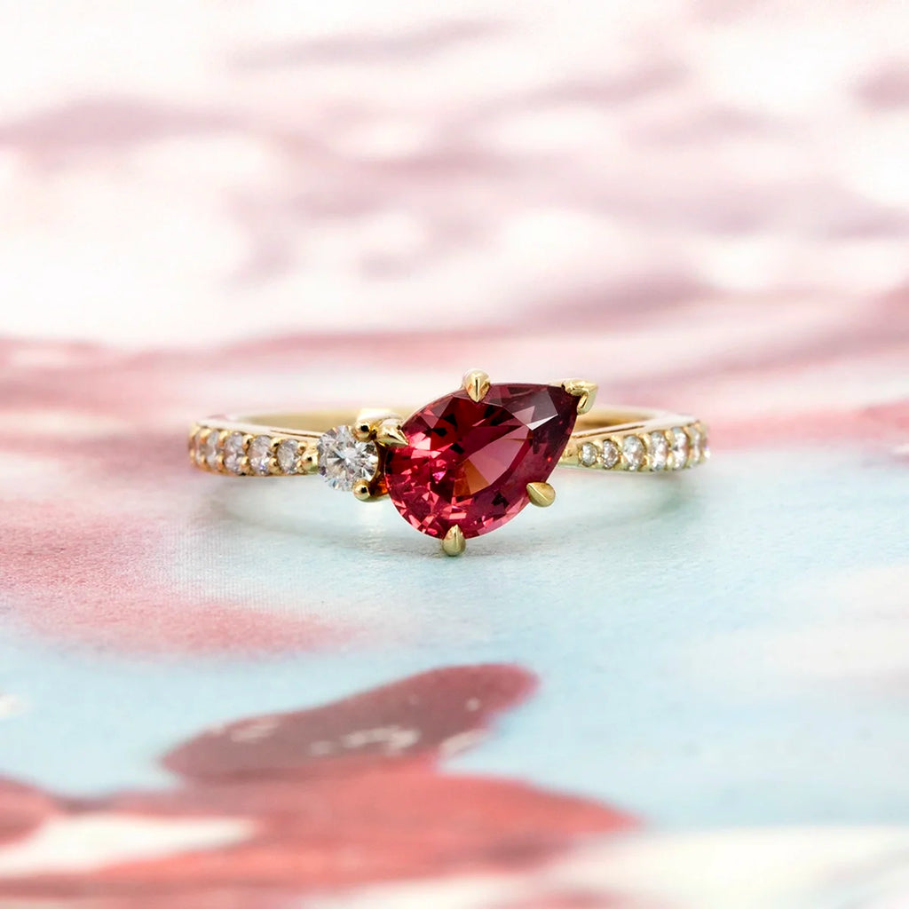 A gold ring handcrafted by Justine Quintal, featuring a pear-shaped raspberry-red spinel gemstone with a round diamond accent and a pavé-set diamond band, displayed on a softly blurred pink and blue background.