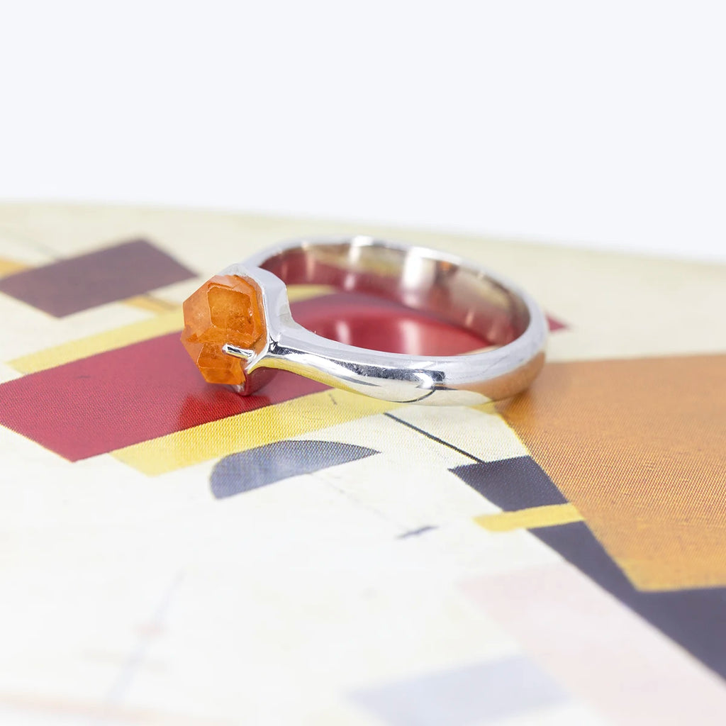 Side view on a funky artistic background of a statement white gold ring featuring a rough orange garnet from Nigeria. This bold piece of jewelry was handcrafted and designed in Montreal by François Charest, and is only available at fine jewelry store Ruby Mardi.