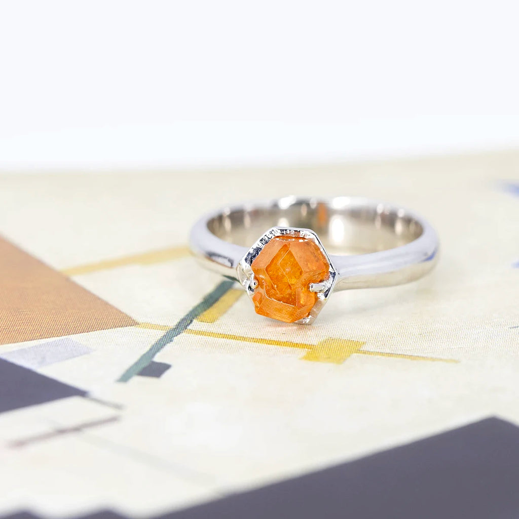 Side view of a white gold statement ring featuring a rough orange gesmtone (spessartite garnet). This gender neutral ring was designed and handcrafted in Montreal by independent jeweller François Charest. An alternative engagement ring or right hand ring seen photographed on an artistic cubic background.