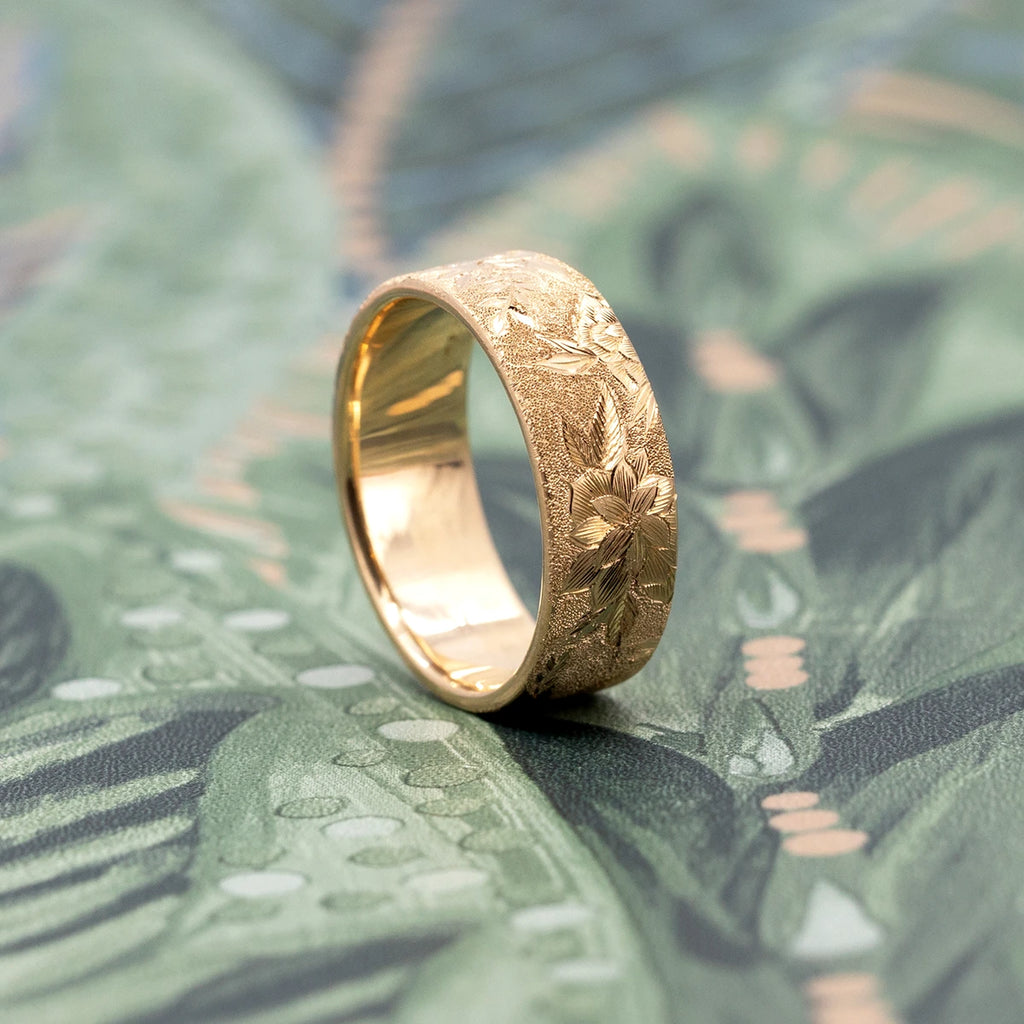 Thick, wide 14-carat yellow gold band featuring finely hand-engraved flowers and a sandblasted texture. The slender jewel is photographed at an angle, against a romantic floral wallpaper. The wedding ring was created by independent brand Rebel & Rogue. This is a unique piece of bridal jewelry only available at Ruby Mardi.
