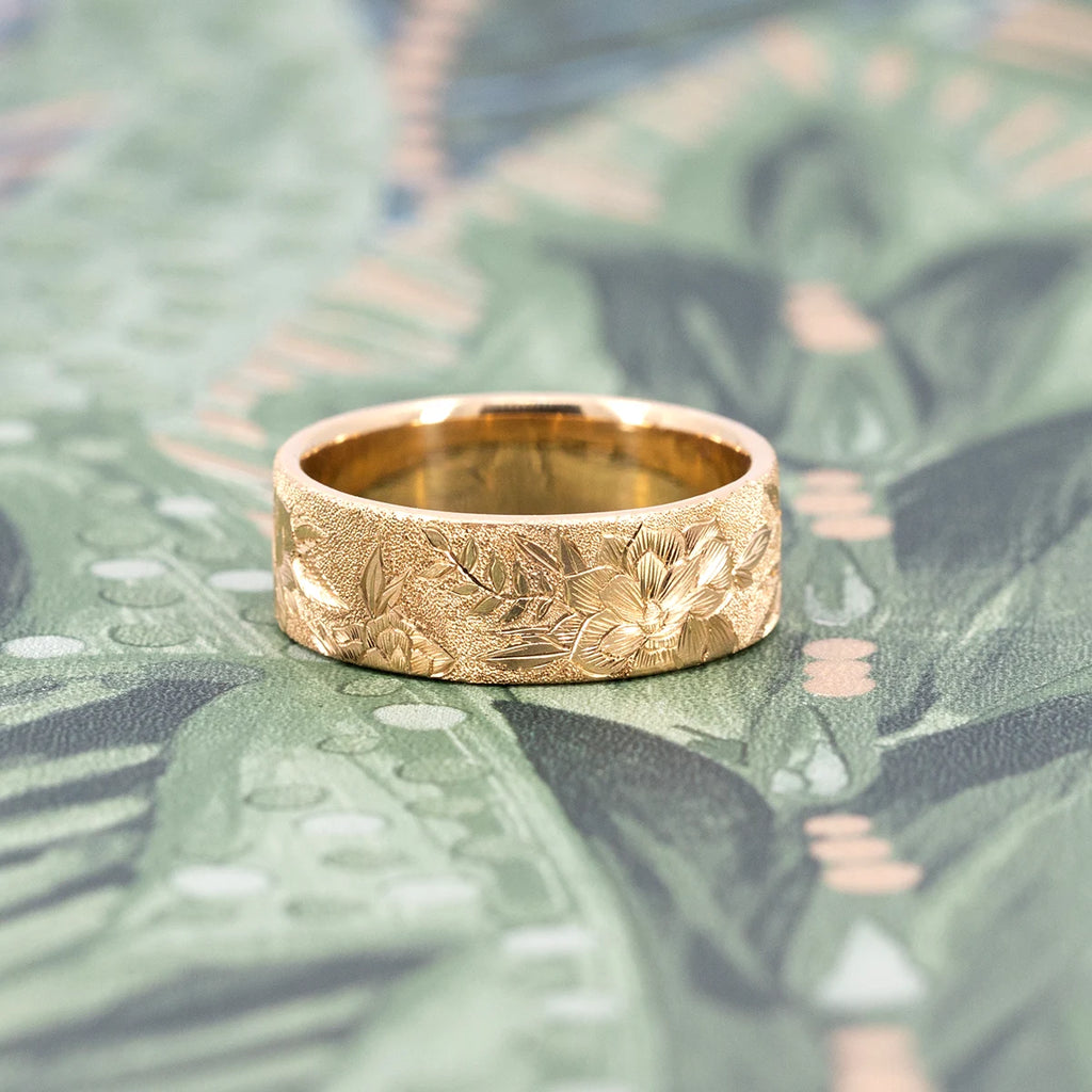 Thick, wide 14-carat yellow gold band featuring finely hand-engraved flowers and a sandblasted texture. The piece of fine jewelry is photographed against a romantic floral wallpaper. The wedding ring was created by independent brand Rebel & Rogue. This is a unique piece of bridal jewelry only available at Ruby Mardi.
