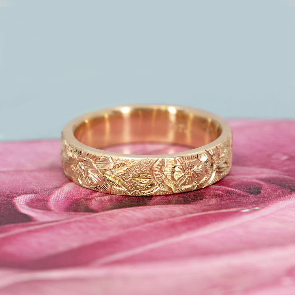 A 14k yellow gold wedding band by Rebel & Rogue resting on a soft pink floral background, showcasing delicate hand-engraved flowers and leaves. The matte-stippled texture contrasts with the polished interior, highlighting the depth of the engraving. Fine jewelry photographed for Ruby Mardi, Montreal.