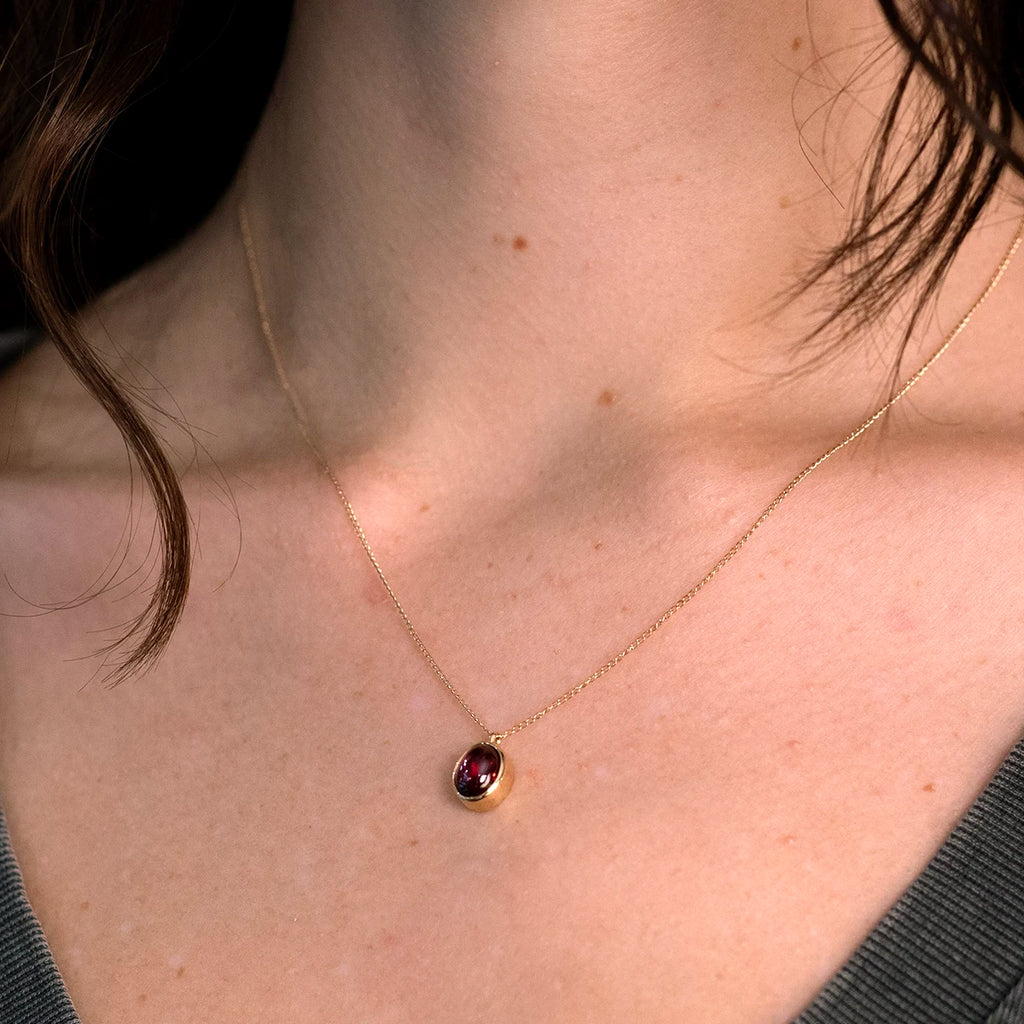A woman wearing a delicate gold chain necklace with an oval red garnet in a bezel setting, handcrafted by Rooz Jewellery and available at Ruby Mardi in Montreal.
