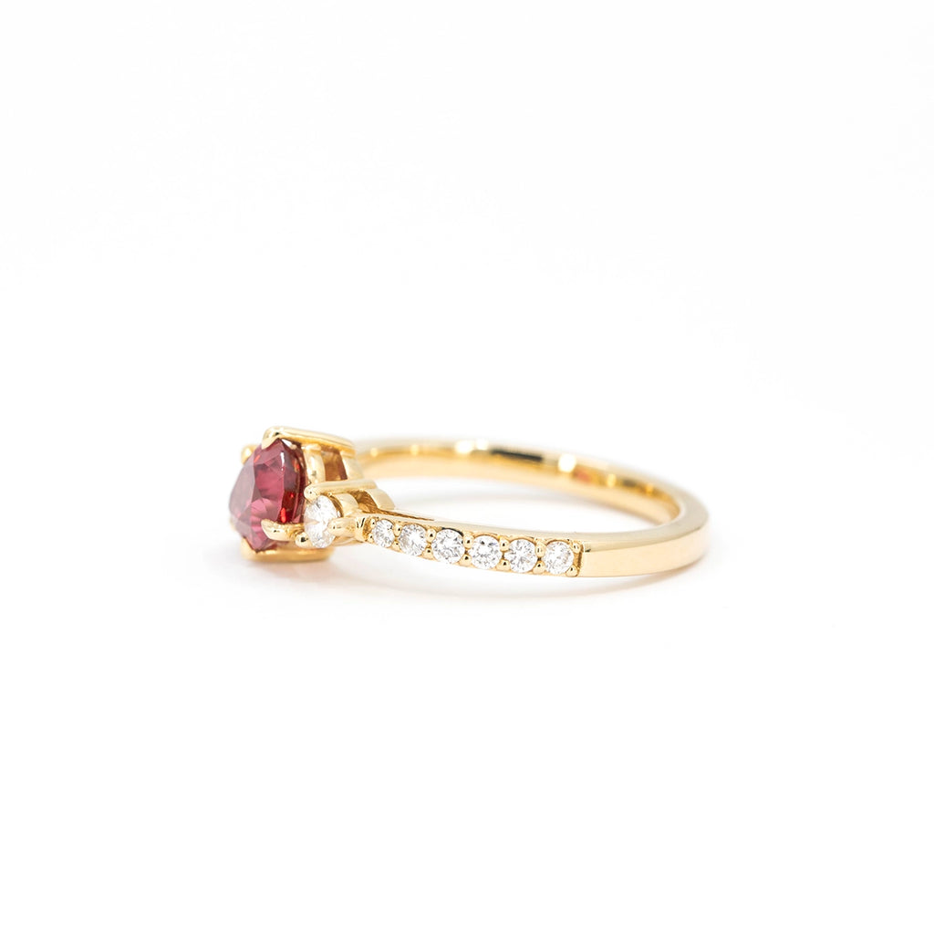Side view of a gold ring handcrafted by Justine Quintal, featuring a pear-shaped red spinel gemstone, a single round diamond accent, and a band adorned with pavé-set diamonds, displayed on a white background.