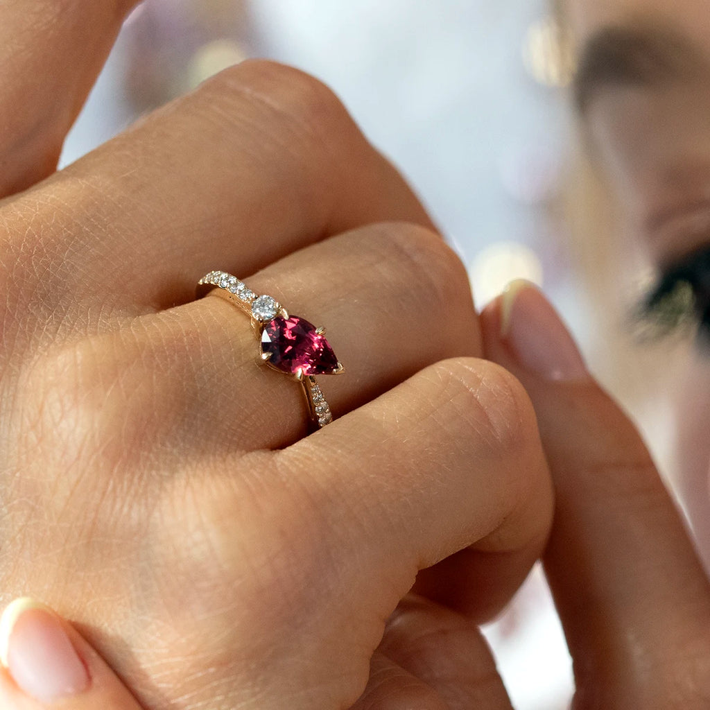 Close-up of a hand wearing a gold ring handcrafted by Justine Quintal, featuring a pear-shaped red spinel gemstone with a round diamond accent and a pavé-set diamond band, with a softly blurred background.